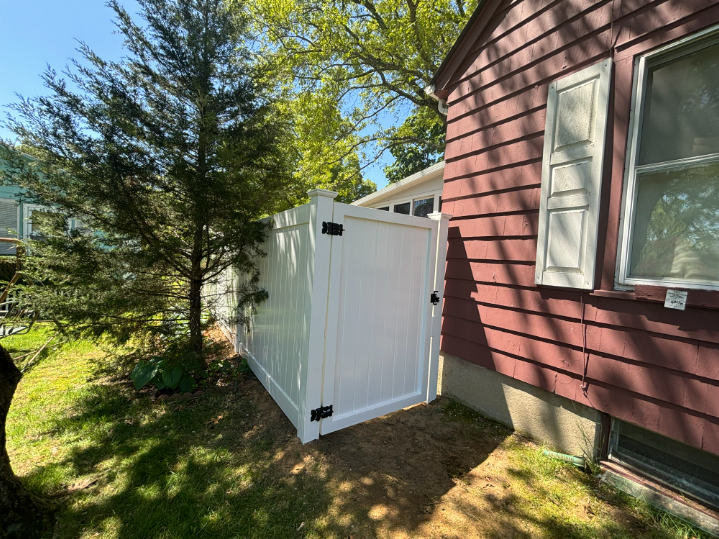 White vinyl fence next to a red house, with a tree on the left side, sunny day.