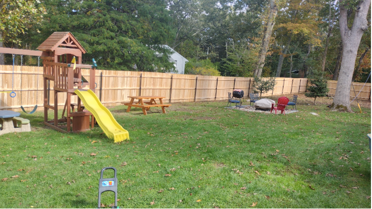 Backyard with wooden playset, picnic table, fire pit, and fence. Green grass and trees.