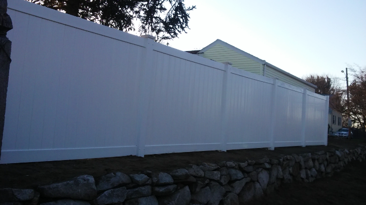 White vinyl fence along a rocky wall, obscuring a house with a green roof against a light sky.