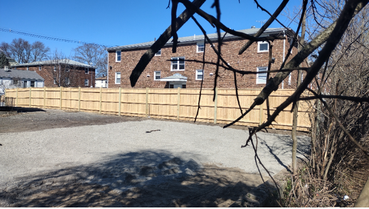 Gravel parking area enclosed by a wooden fence, with a brick apartment building in the background on a sunny day.