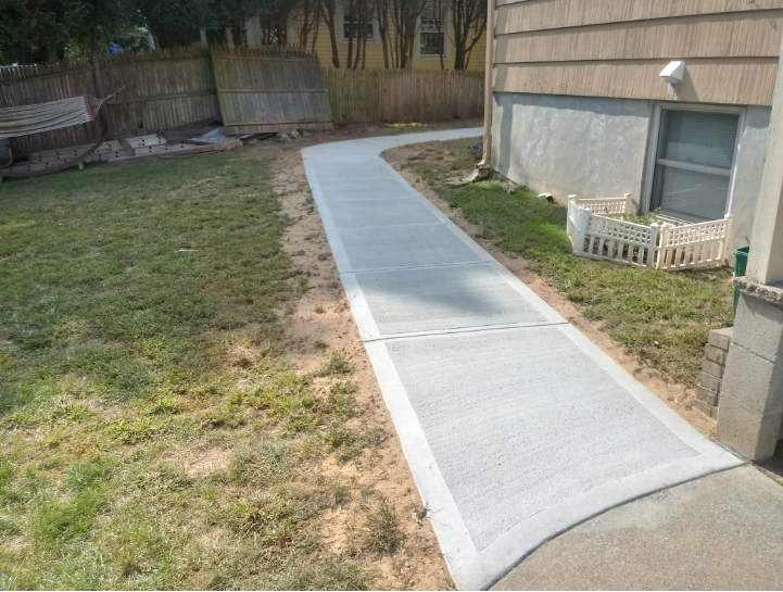 Concrete walkway curving through a grassy backyard, adjacent to a house with beige siding.