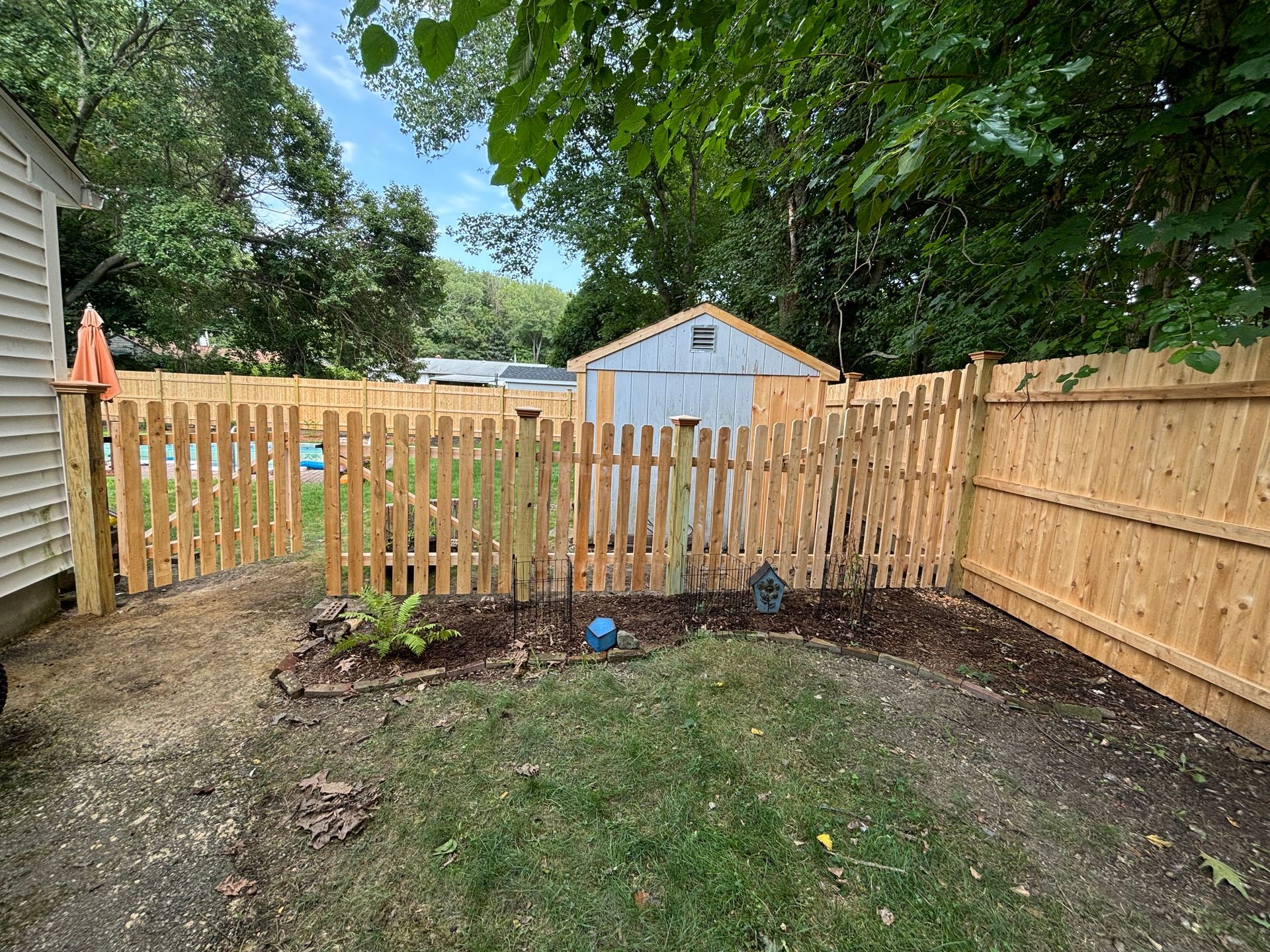 A wooden picket fence encloses a backyard with a garden, shed, and trees.