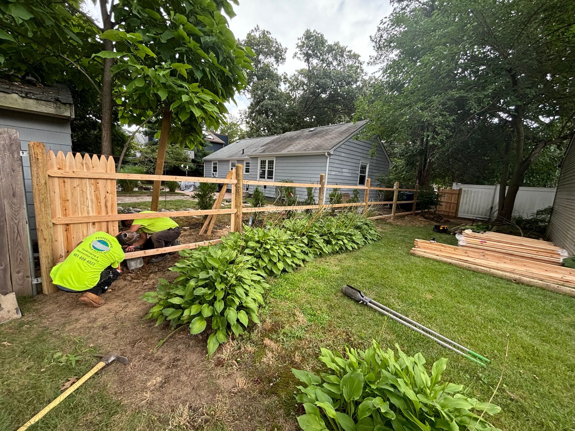 A person installing a wooden fence in a backyard, with a row of green plants and a house in the background.