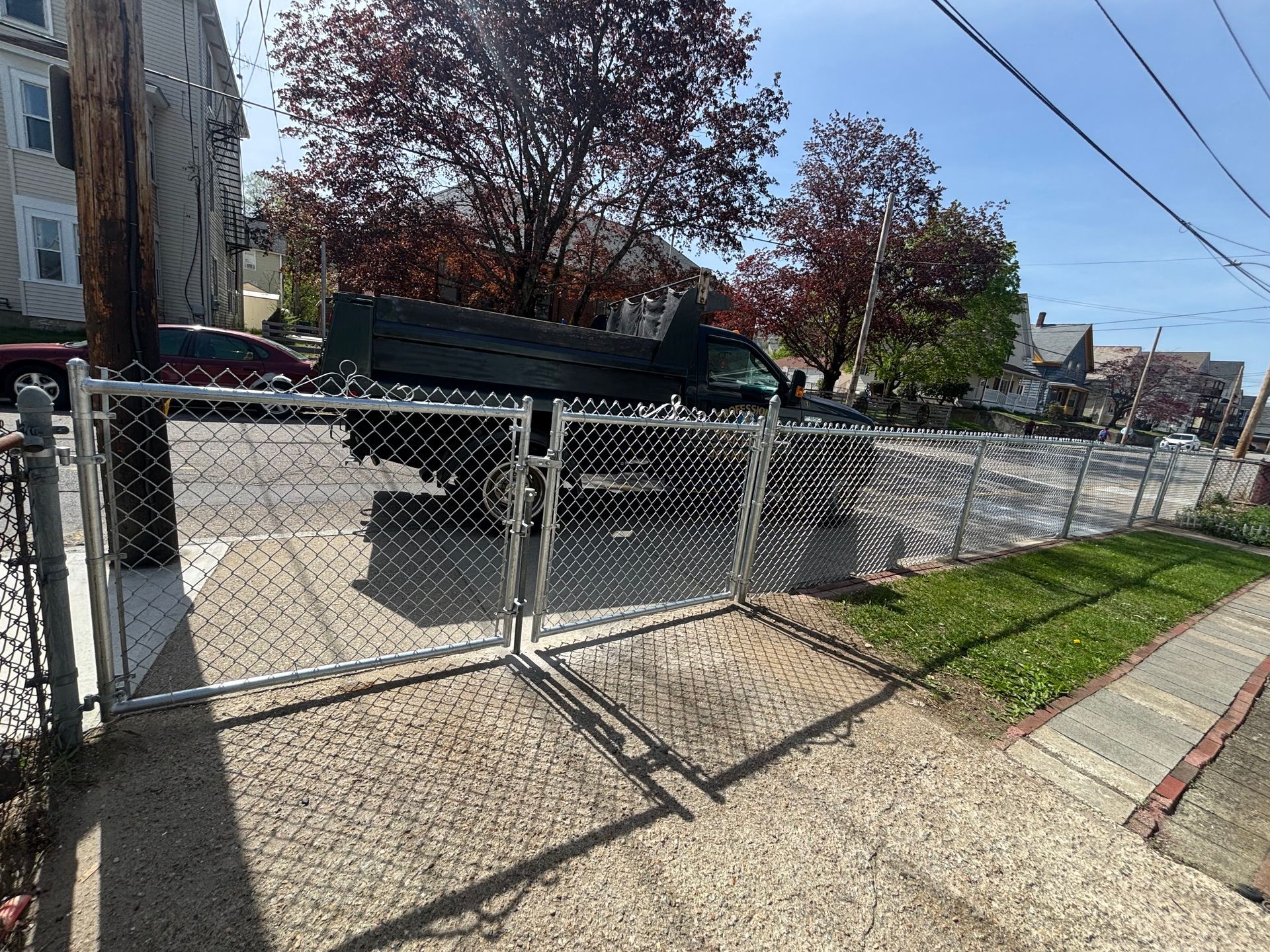 Chain-link fence with gate beside a sidewalk, a truck parked behind it, and houses in the background.