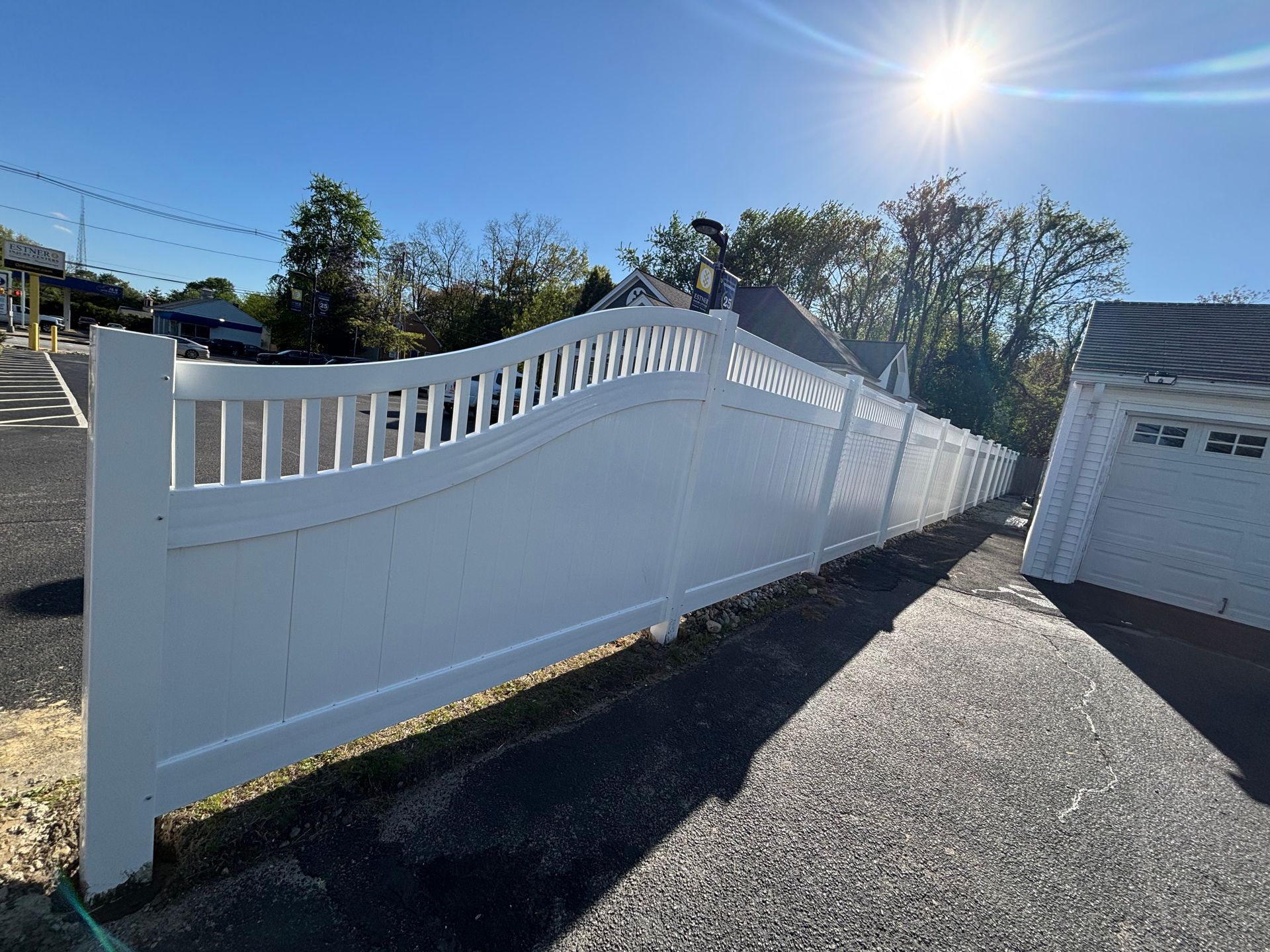 White vinyl fence curves along a paved driveway under a bright sun.