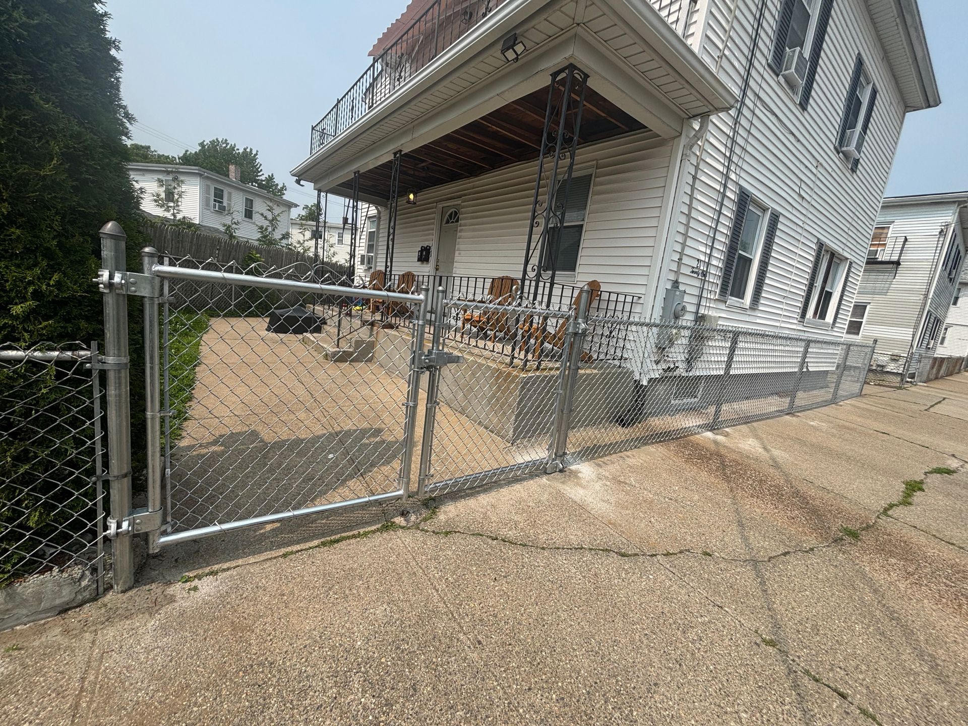 Chain-link fence and gate in front of a white house with a porch and concrete driveway.