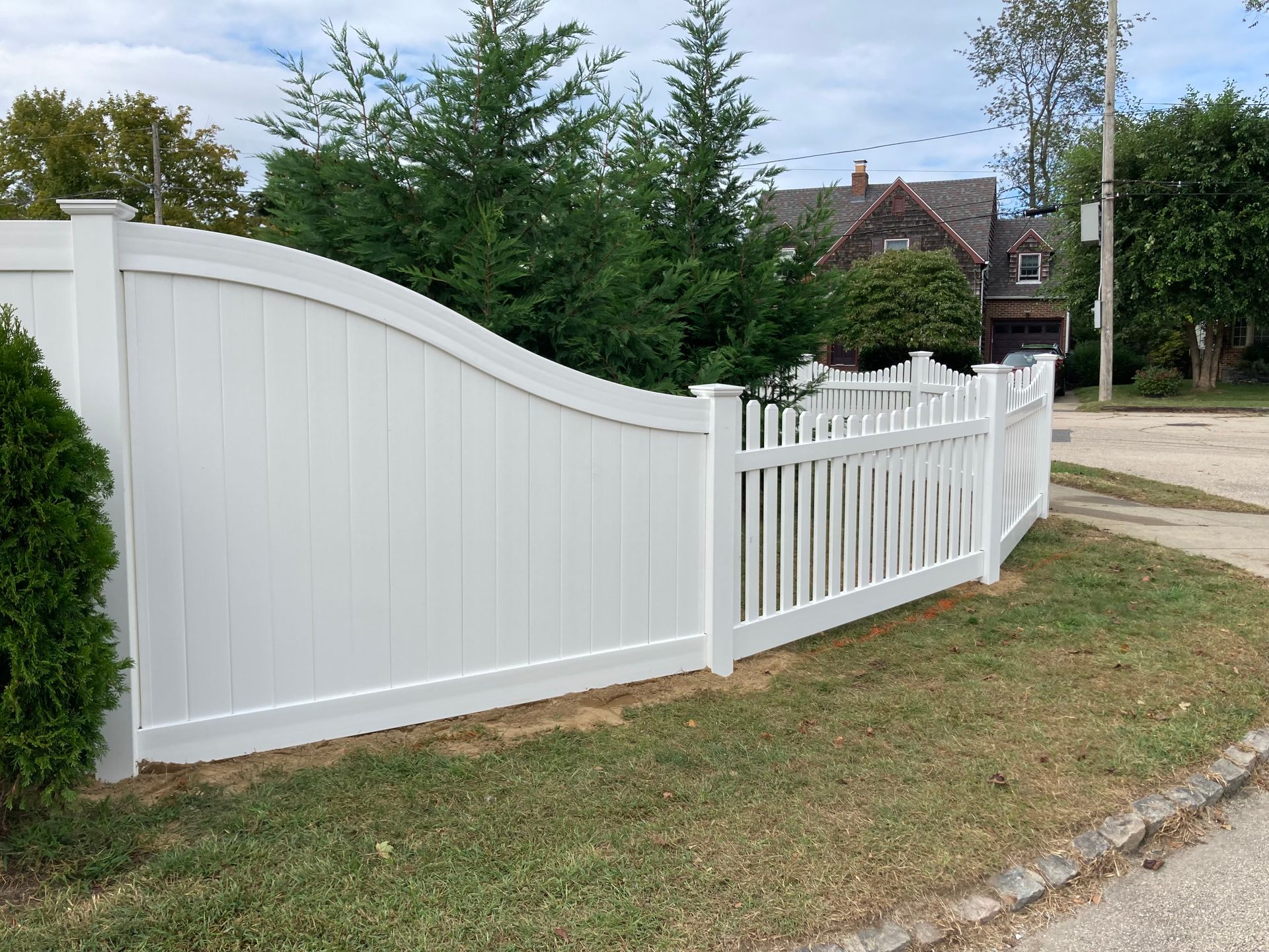 White vinyl fence curves around a yard, with green bushes and a house in the background.