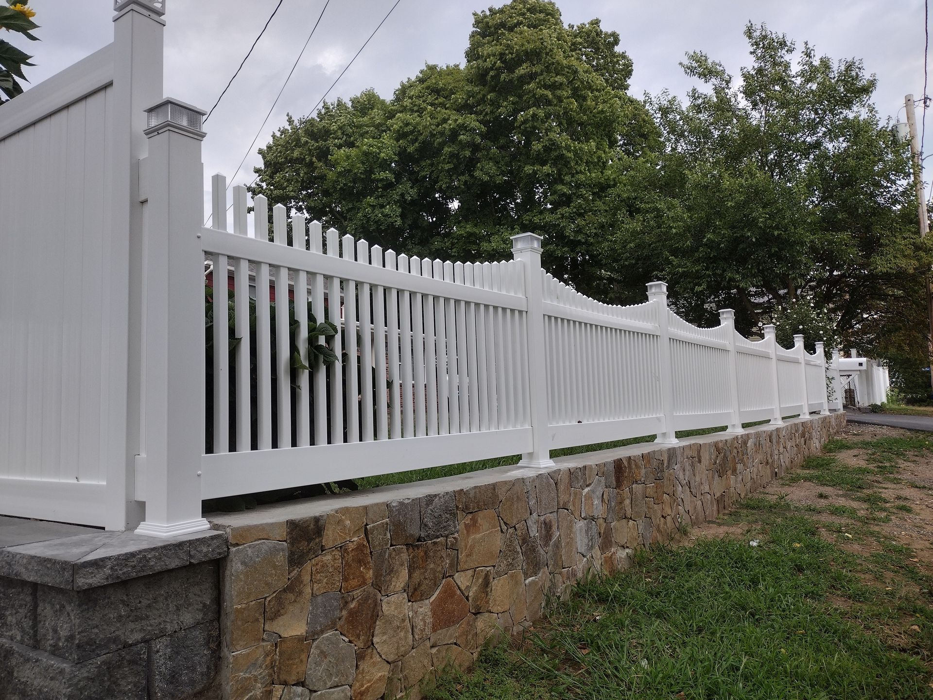 White picket fence atop a stone wall, bordering a grassy area and street.