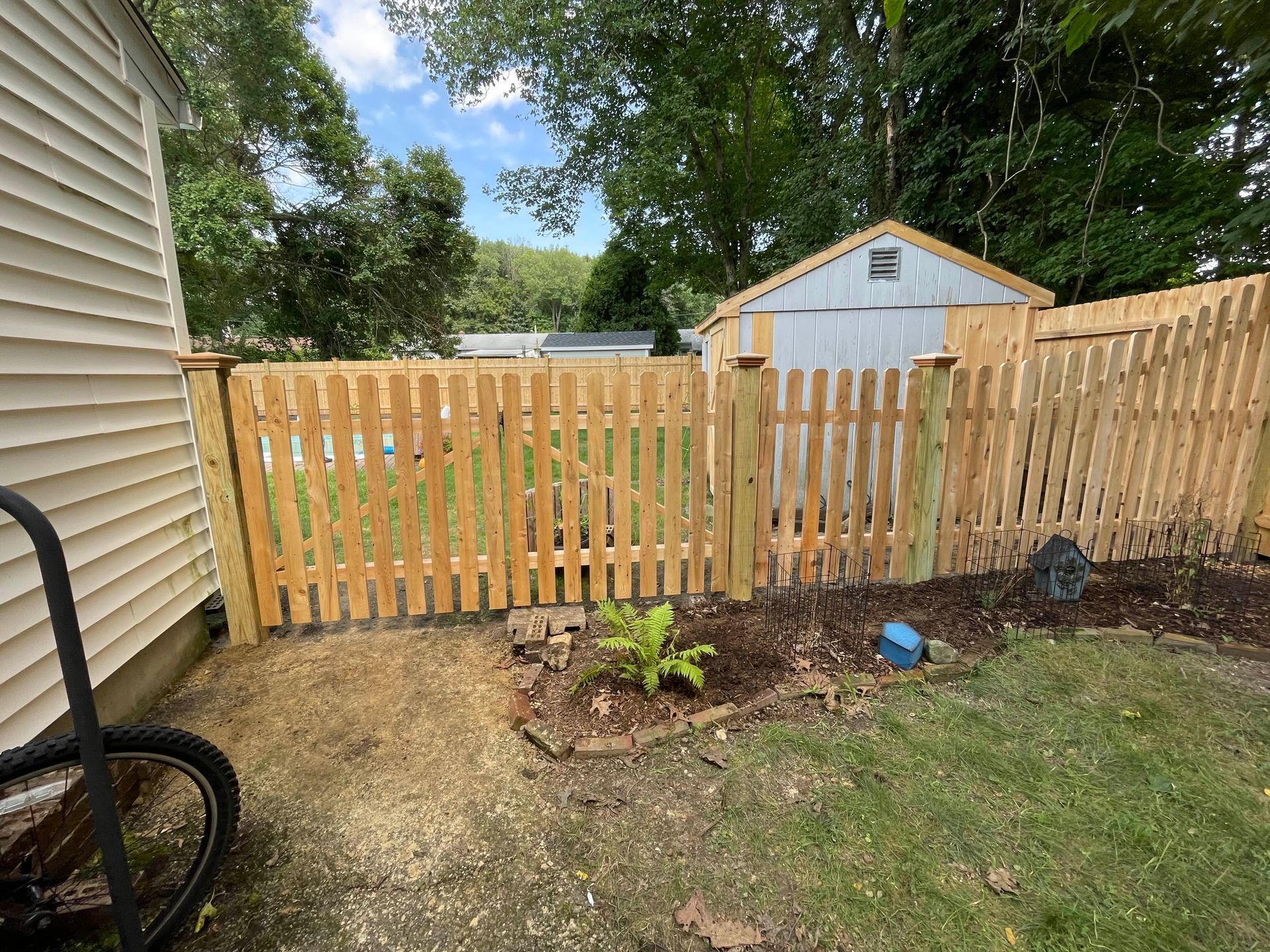 Wooden picket fence surrounds a yard with a shed in the background, next to a house.