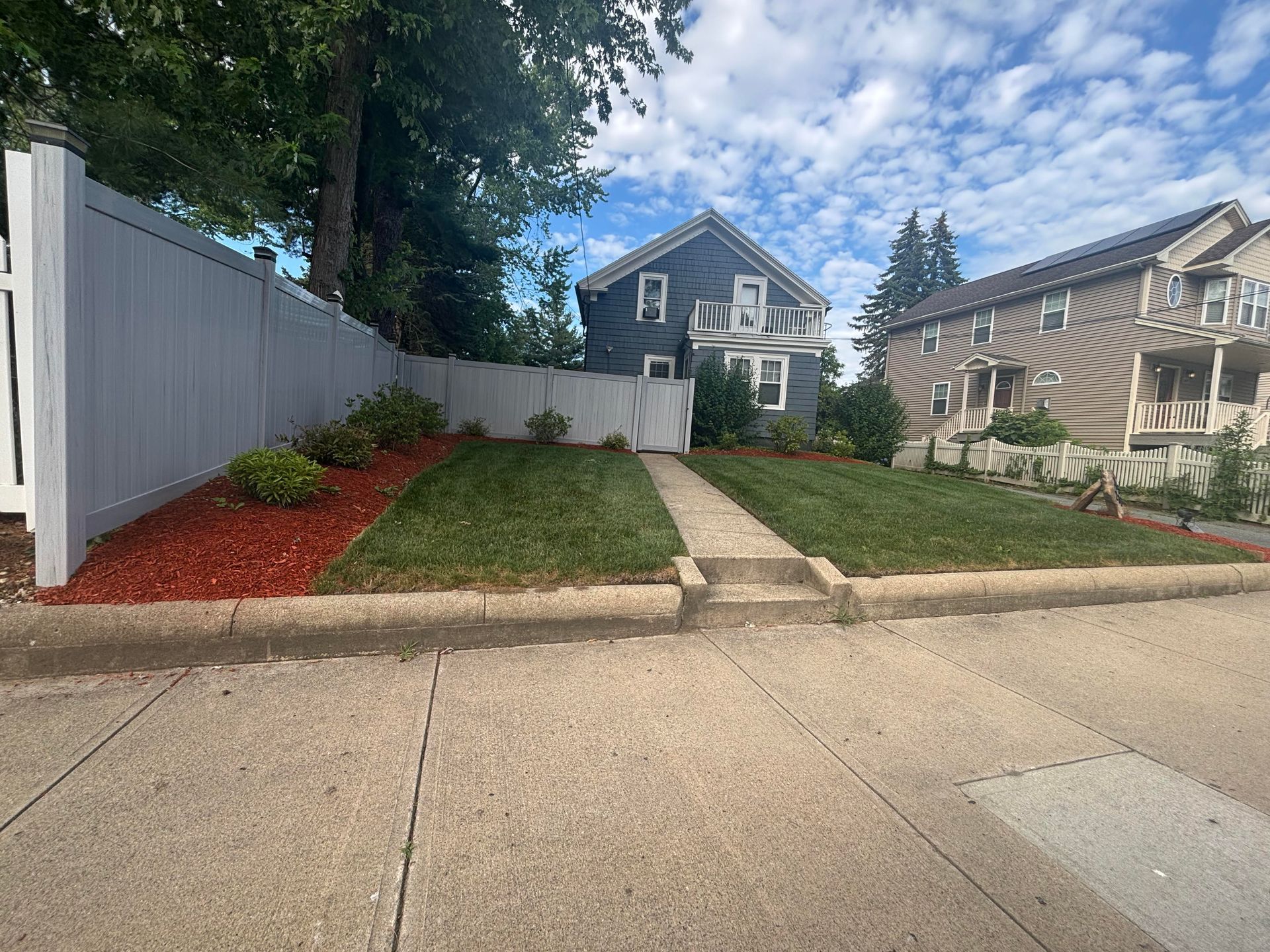 A house with a gray exterior, a small lawn, and a gray fence, with a sidewalk in front.