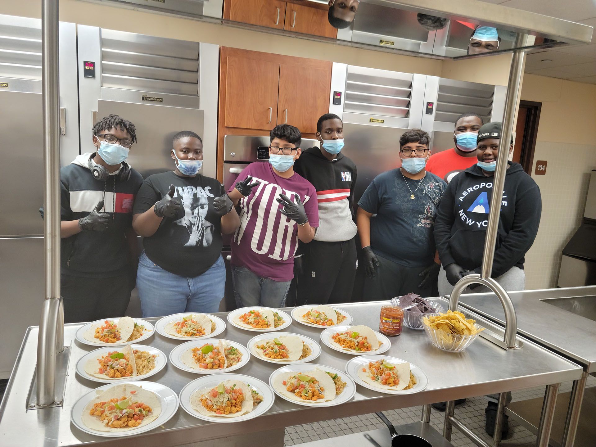 Group prepares plates of food in a kitchen. Several people wear masks and gloves, with prepared tacos on the counter.