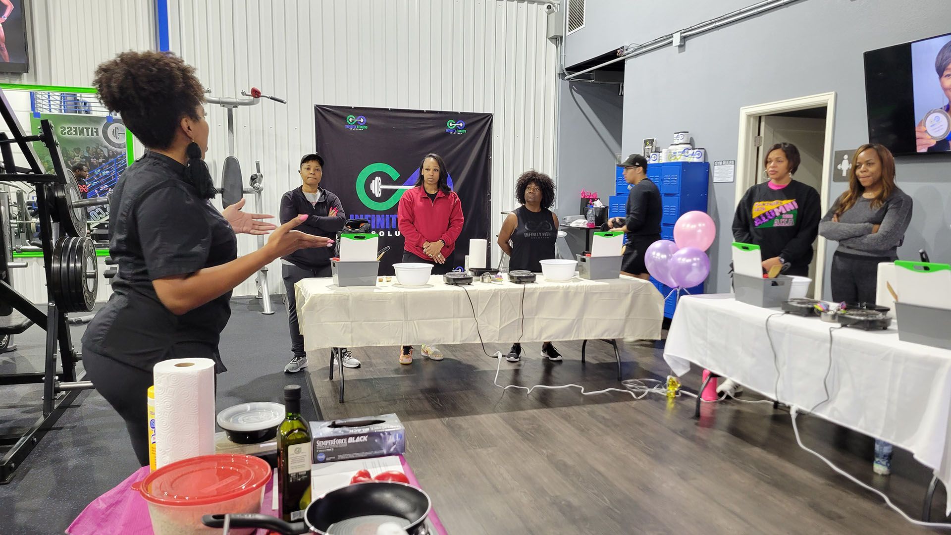 Woman speaking to a panel at a cooking event in a gym. Attendees stand behind tables.