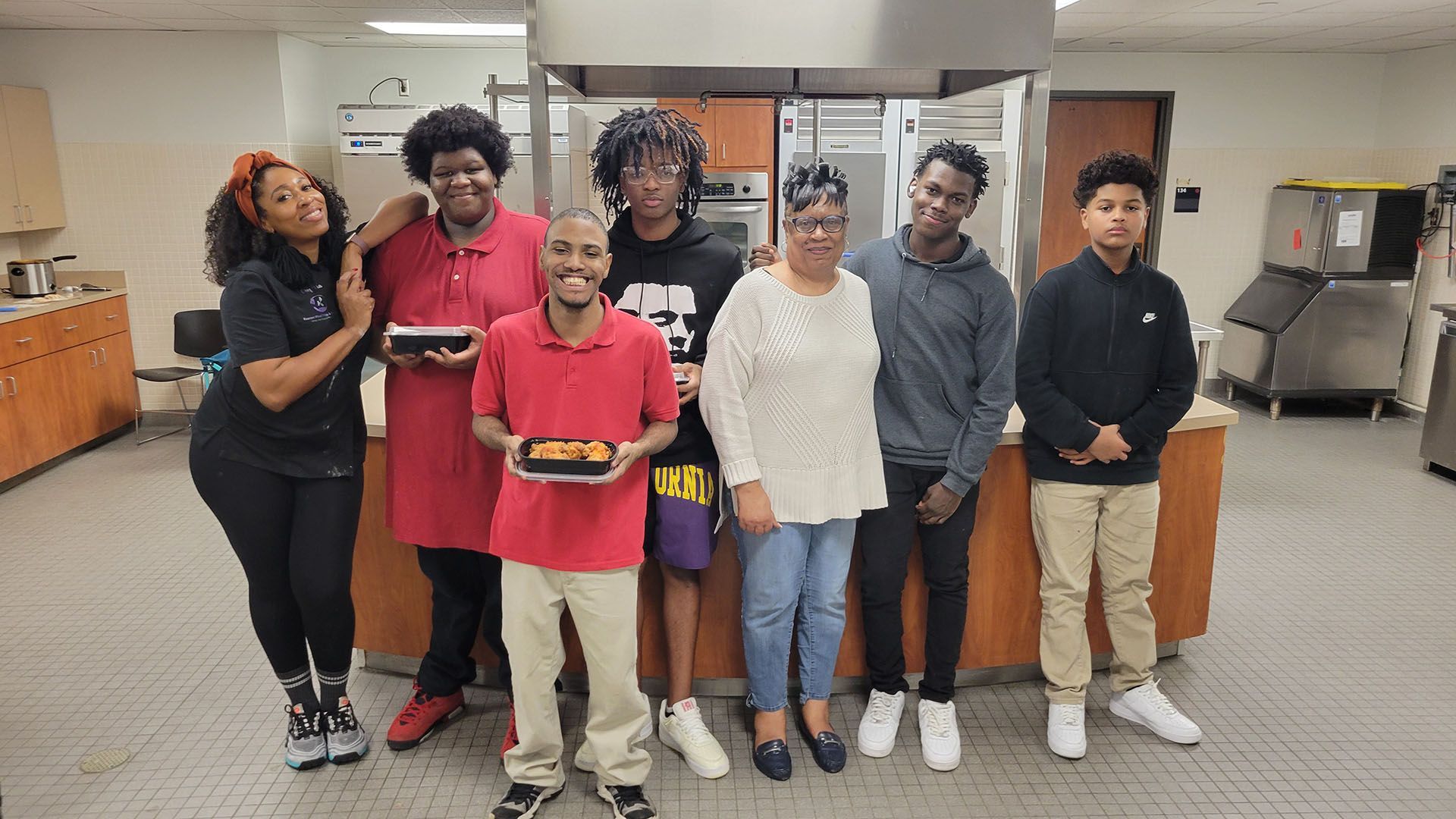 Group of people in a kitchen holding trays of food, smiling.