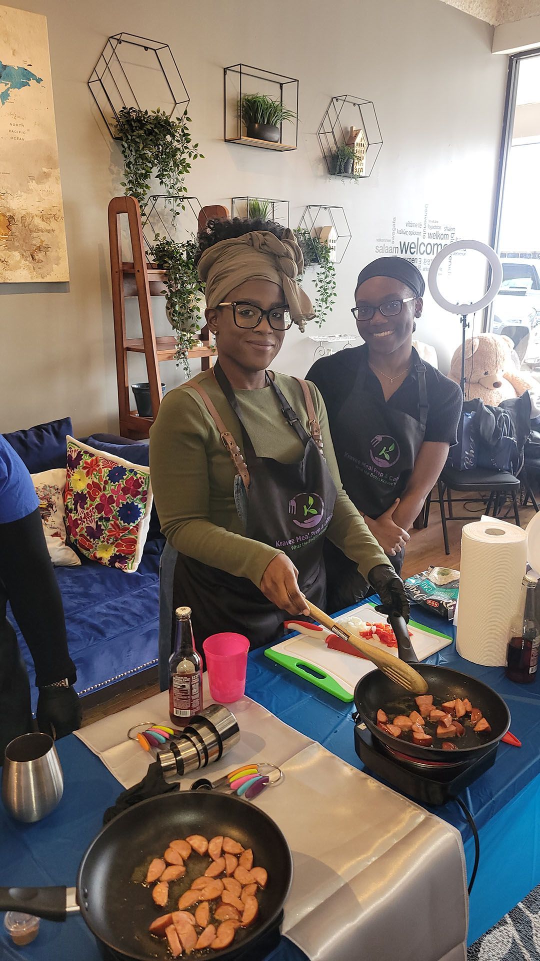 Two people cooking at a demonstration. One person stirs food in a pan, and another smiles. Plants decorate the wall.