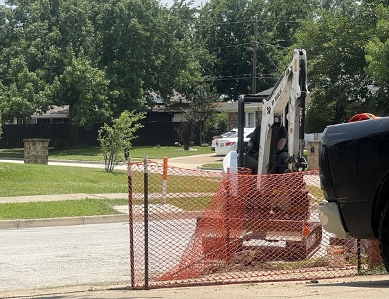 Backhoe behind orange safety fence on a street, with trees and grass in the background