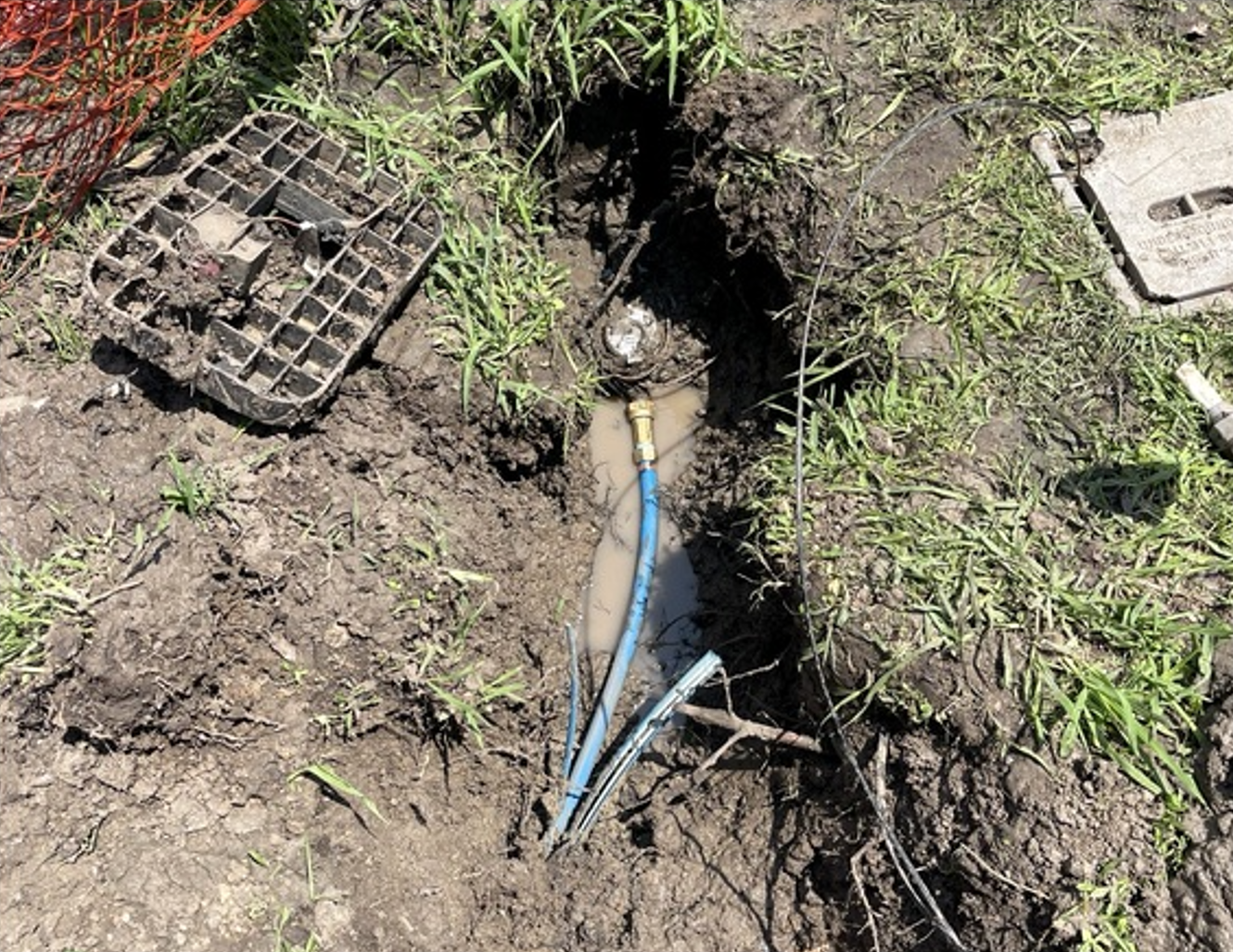 Broken blue pipe leaking into a muddy hole beside grass and a drain grate