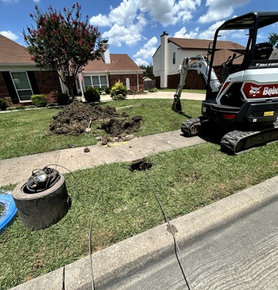 Small excavator near a house digs up lawn beside sidewalk on a sunny suburban street
