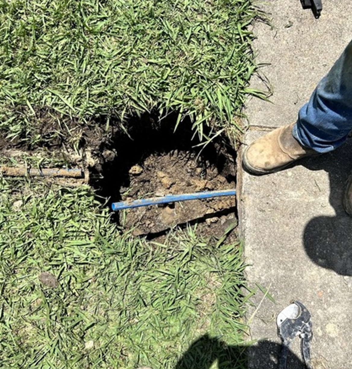 A sinkhole beside a sidewalk with exposed dirt, a blue pipe, and two people’s shoes nearby.