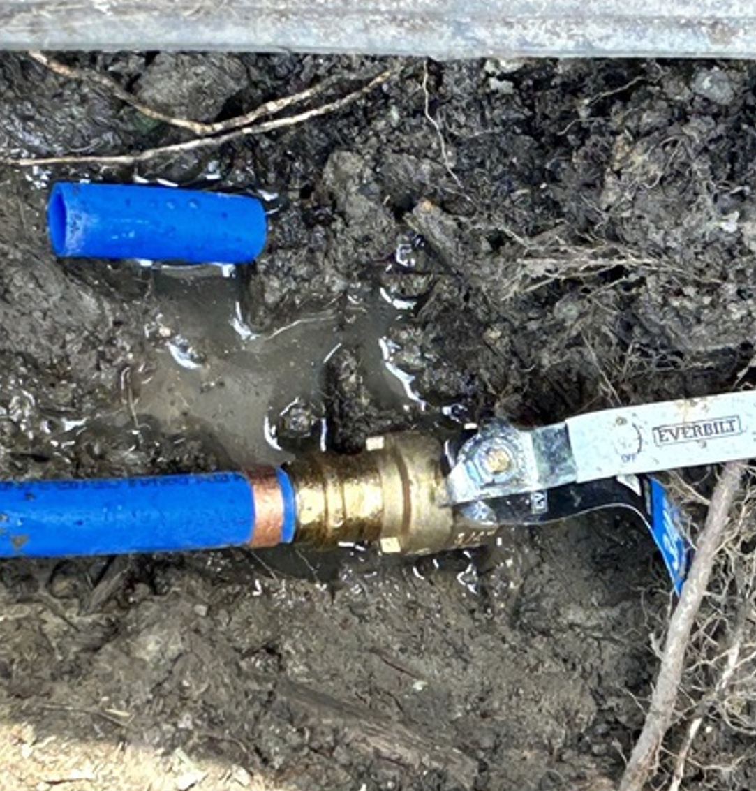 Blue pipe connected to a brass valve in muddy ground, with a wrench attached nearby.