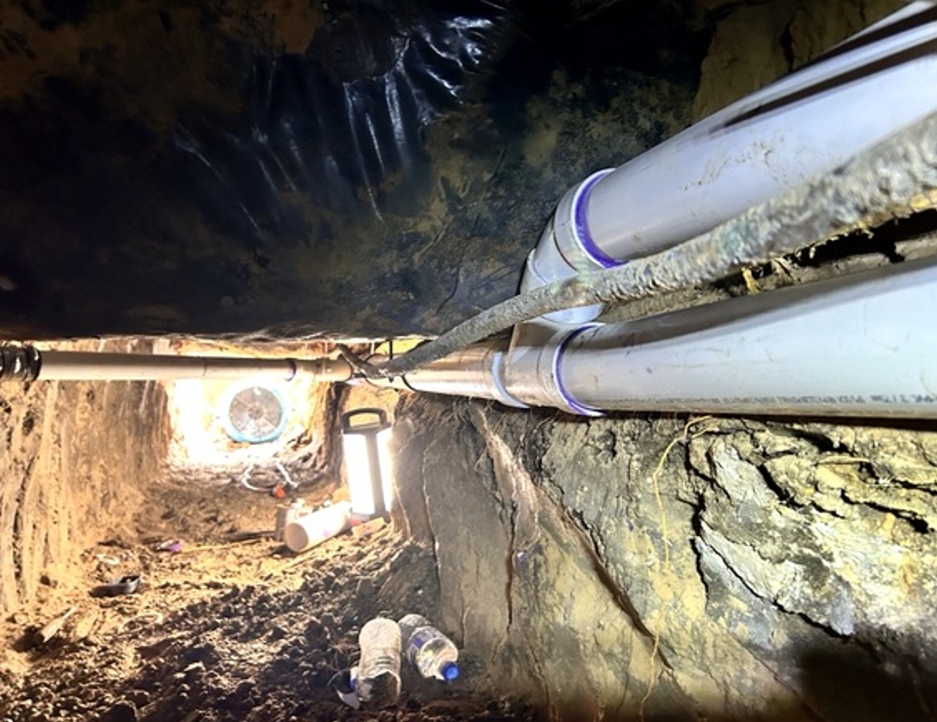 Crawlspace with exposed pipes along a muddy trench and a bright light at the far end.