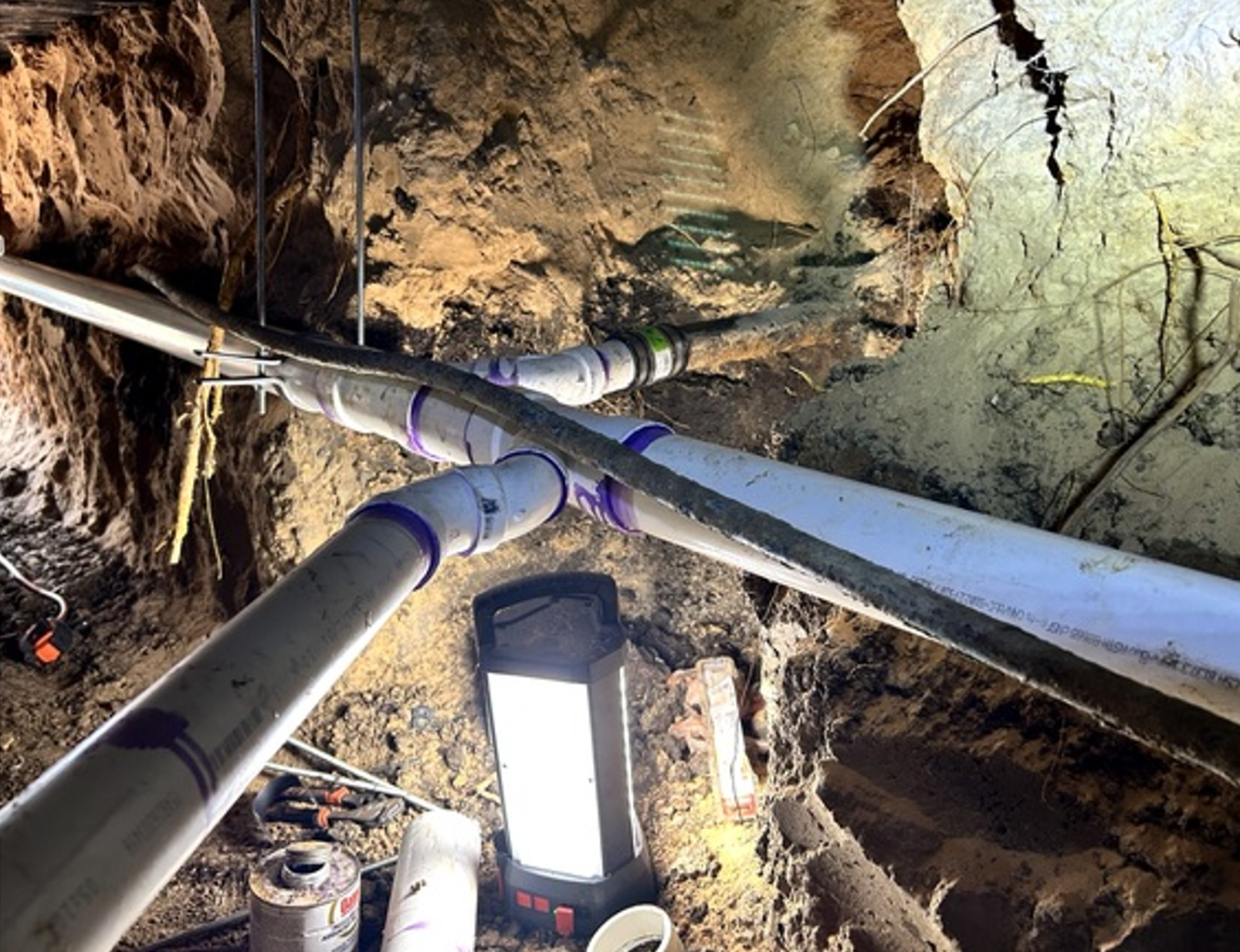 Underground utility pipes and a bright work light inside a rough dirt excavation tunnel