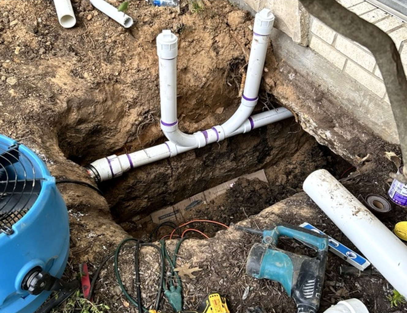 Trenched PVC plumbing pipes and fittings beside a brick wall and blue equipment at a construction site