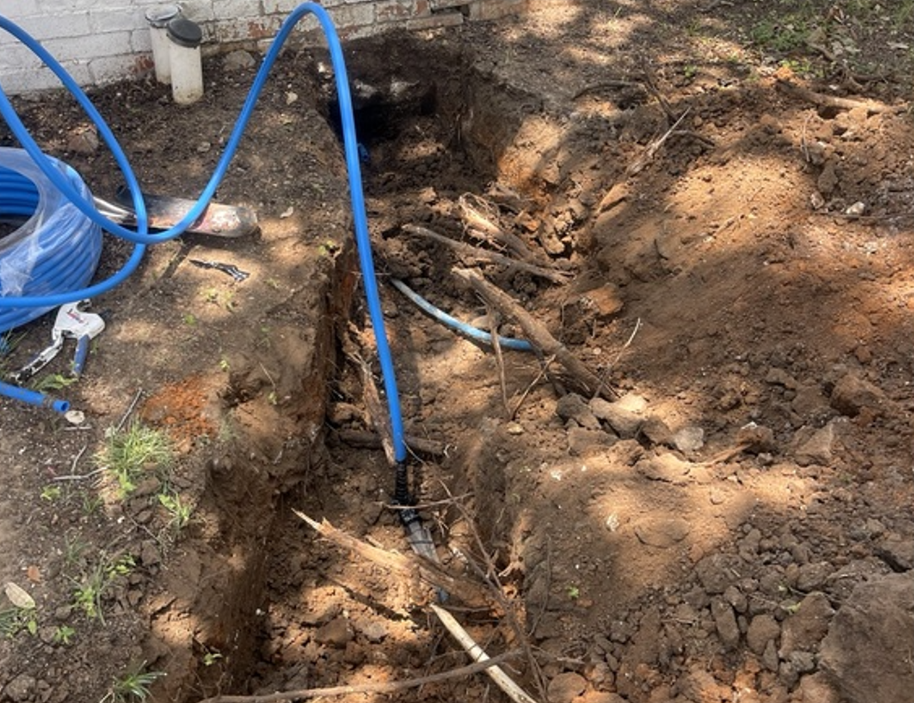Blue hose on dirt and rocks, with a trench and exposed pipes in a work area.