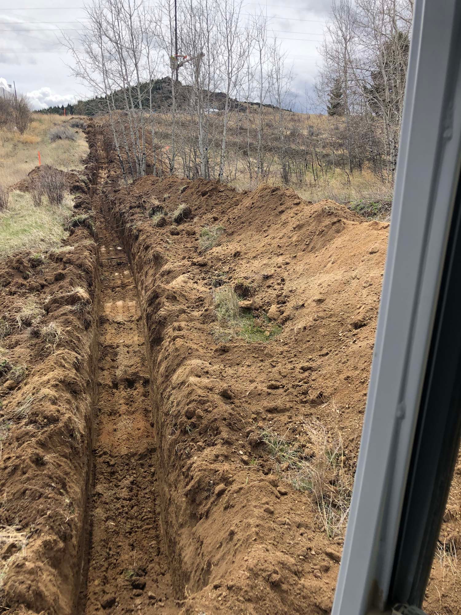 Deep trench cut through brown dirt in a field, viewed from a doorway with bare trees in the background