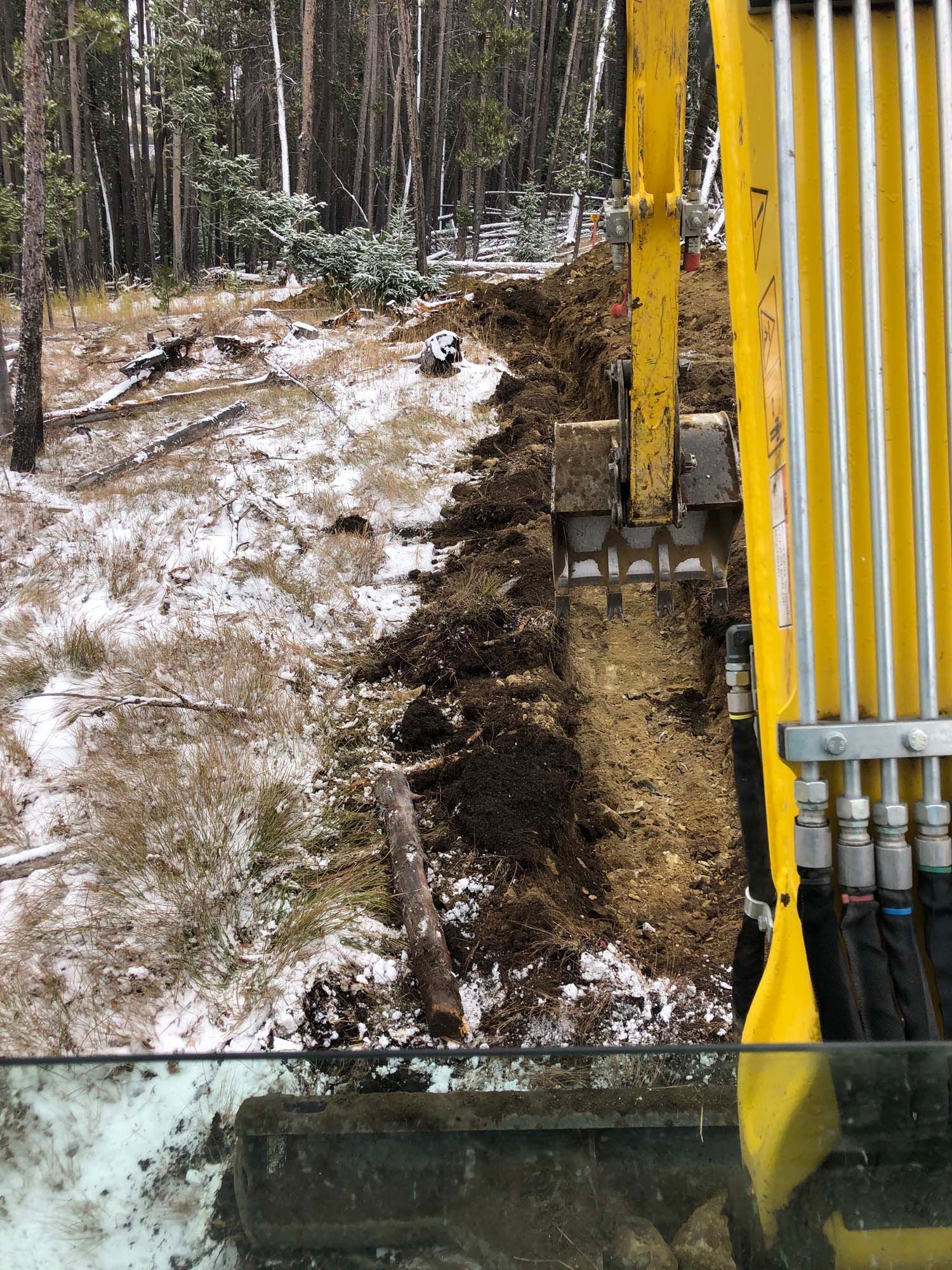 Yellow excavator arm beside a muddy, snow-covered forest path and a shallow ditch