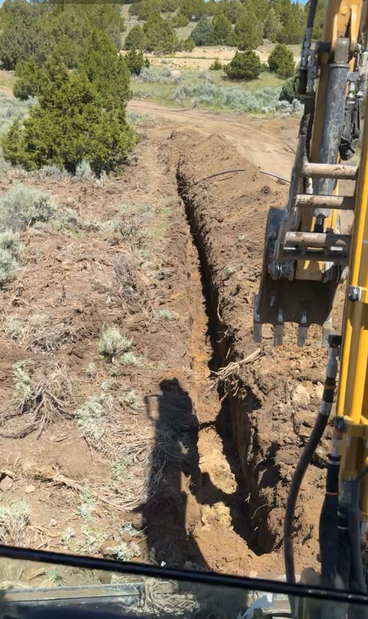 Backhoe digging a trench along a dirt path in a rural, wooded area.