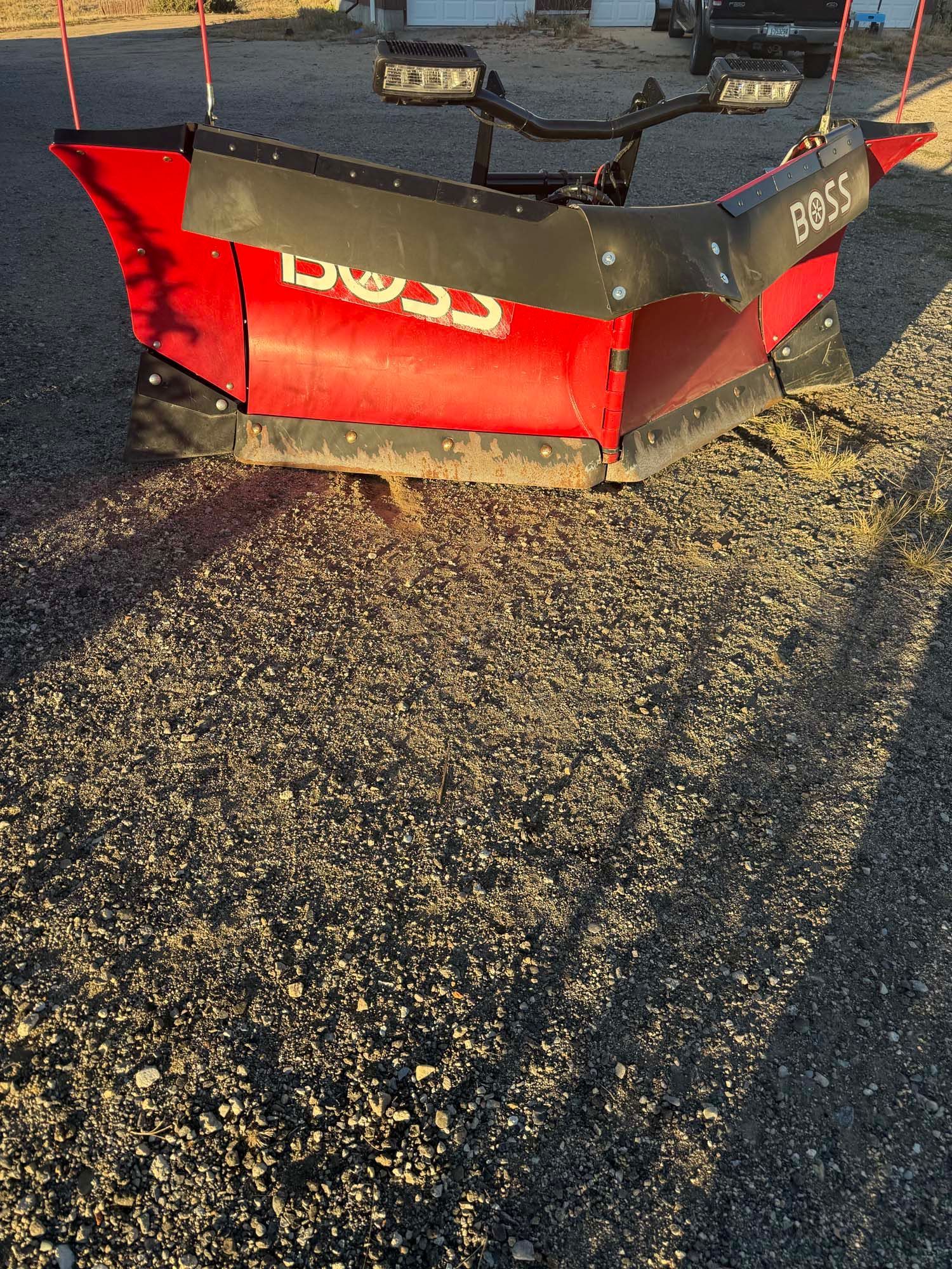 Red snowplow blade attached to a vehicle, parked on gravel in bright sunlight.