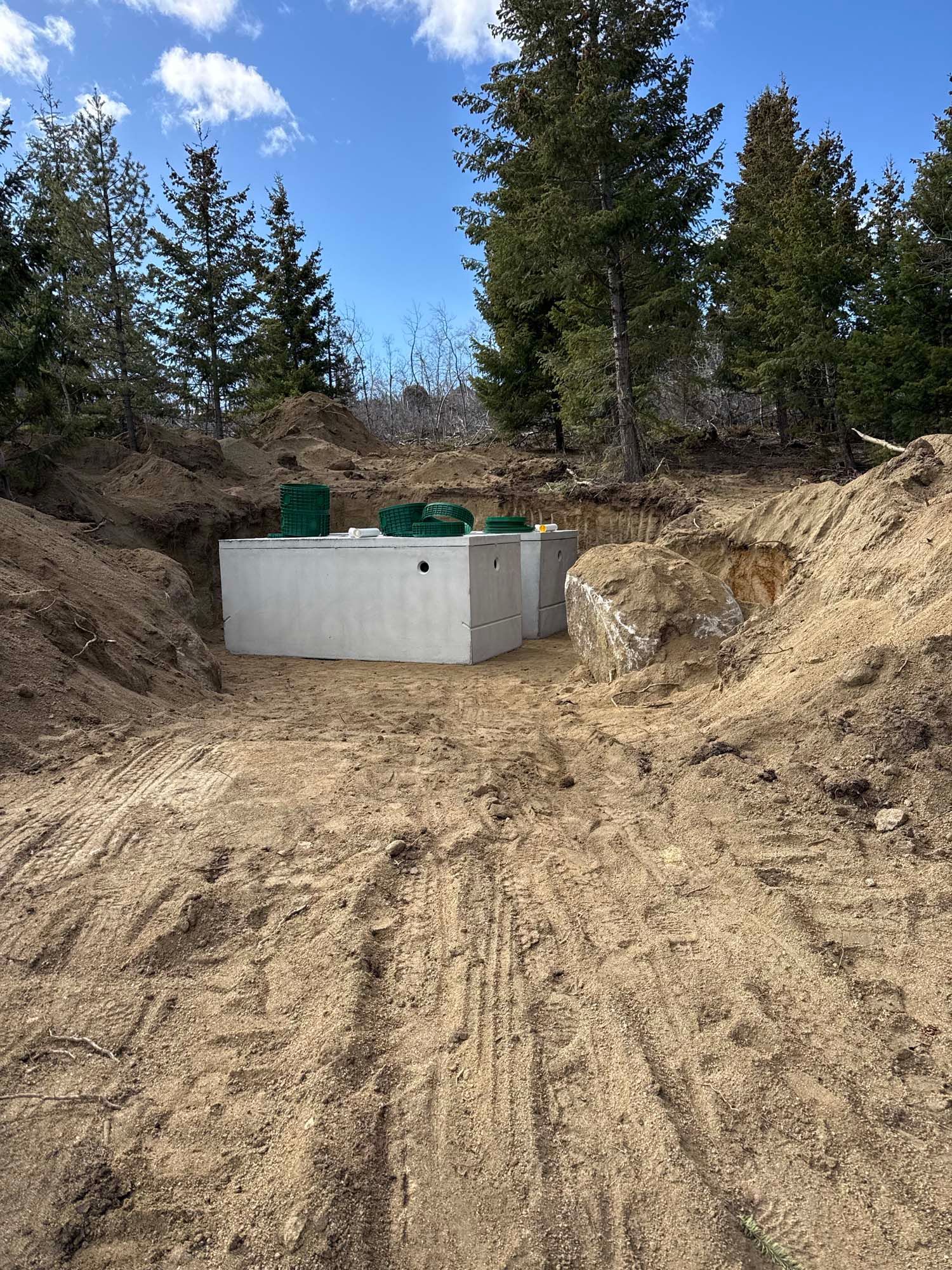 Concrete utility tanks in a dirt trench surrounded by pine trees under a blue sky