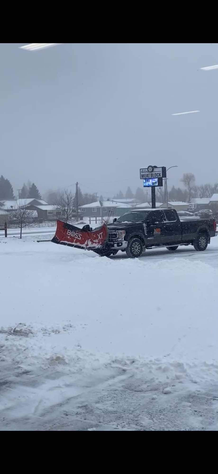 Pickup truck with a red snowplow pushing snow in a snowy parking lot during a snowfall