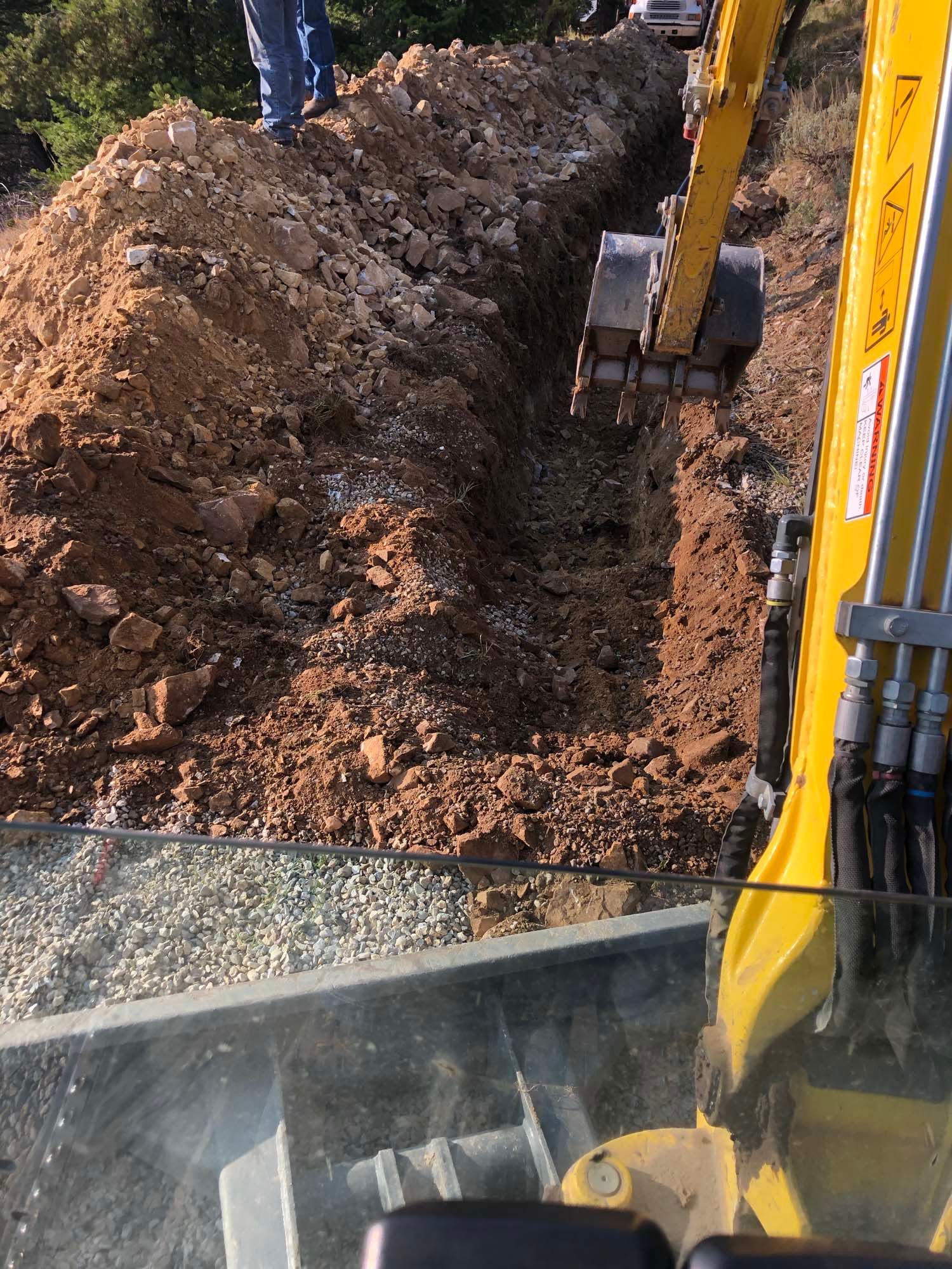 Excavator digging a trench beside a dirt embankment, with soil piled along one side.