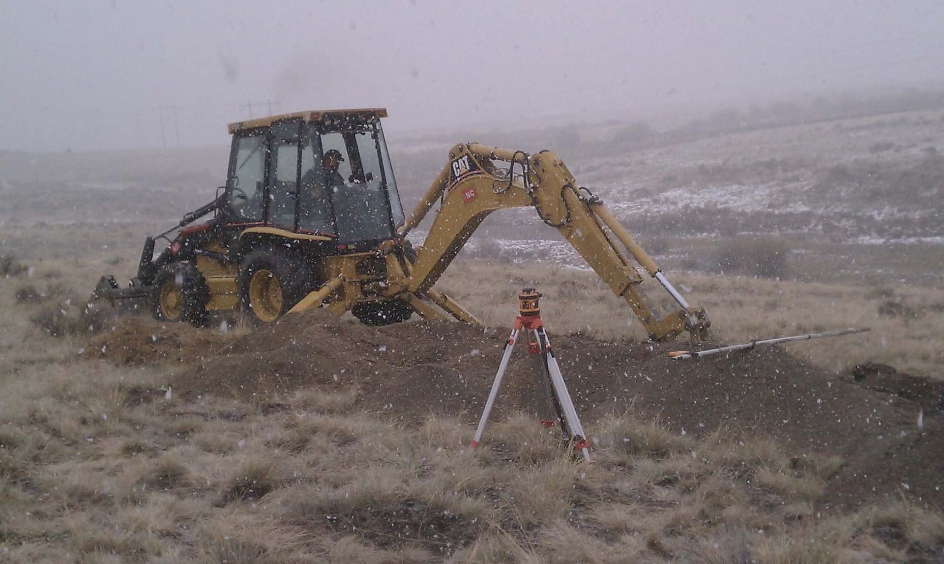 Backhoe excavator digging in a snowy field beside a survey tripod