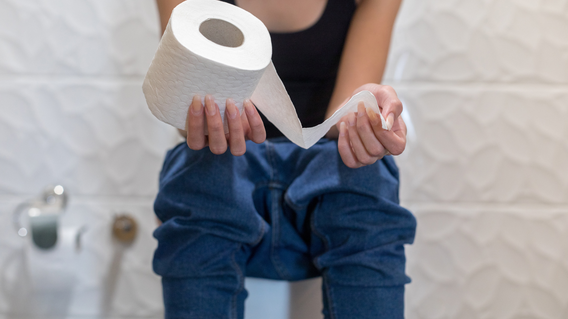 Person sitting on a toilet, holding a roll of toilet paper in a bathroom.