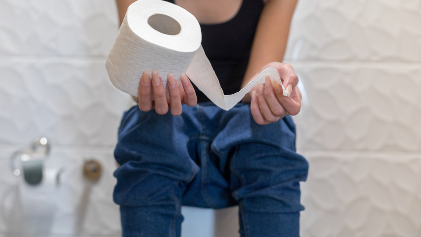 Person sitting on a toilet, holding a roll of toilet paper in a bathroom.