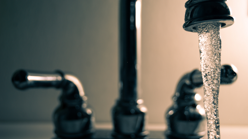 Water flowing from a kitchen faucet over a sink, with a blurred second faucet in the background