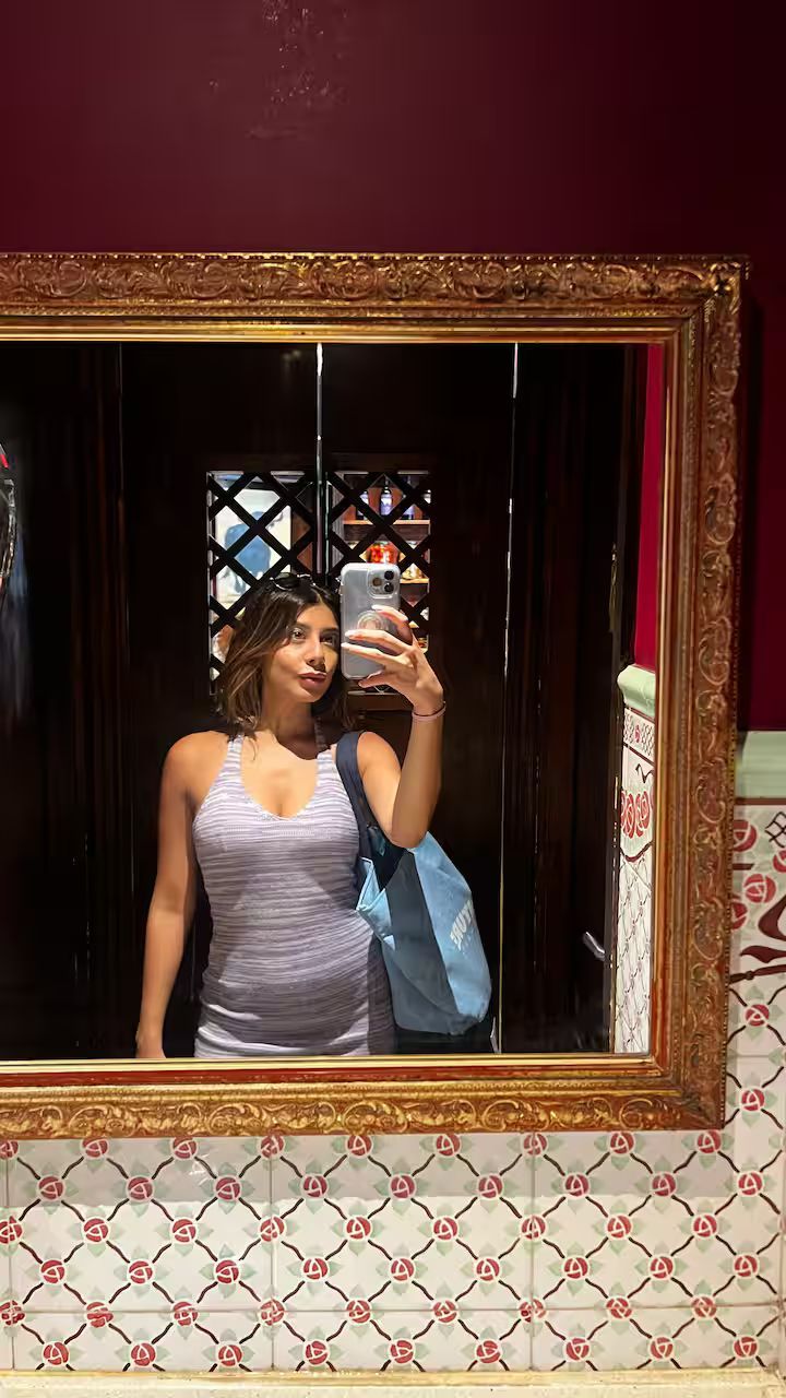 Woman taking a mirror selfie in a framed wall mirror, wearing a lilac dress in a patterned room