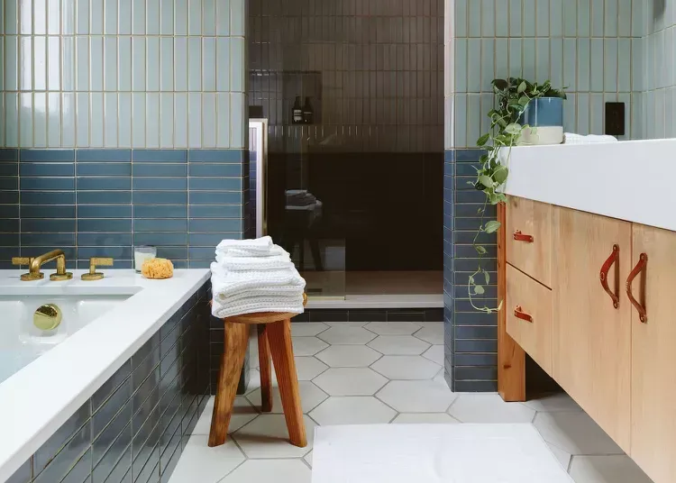 A bathroom with blue tiles , a wooden stool , a sink and a bathtub.