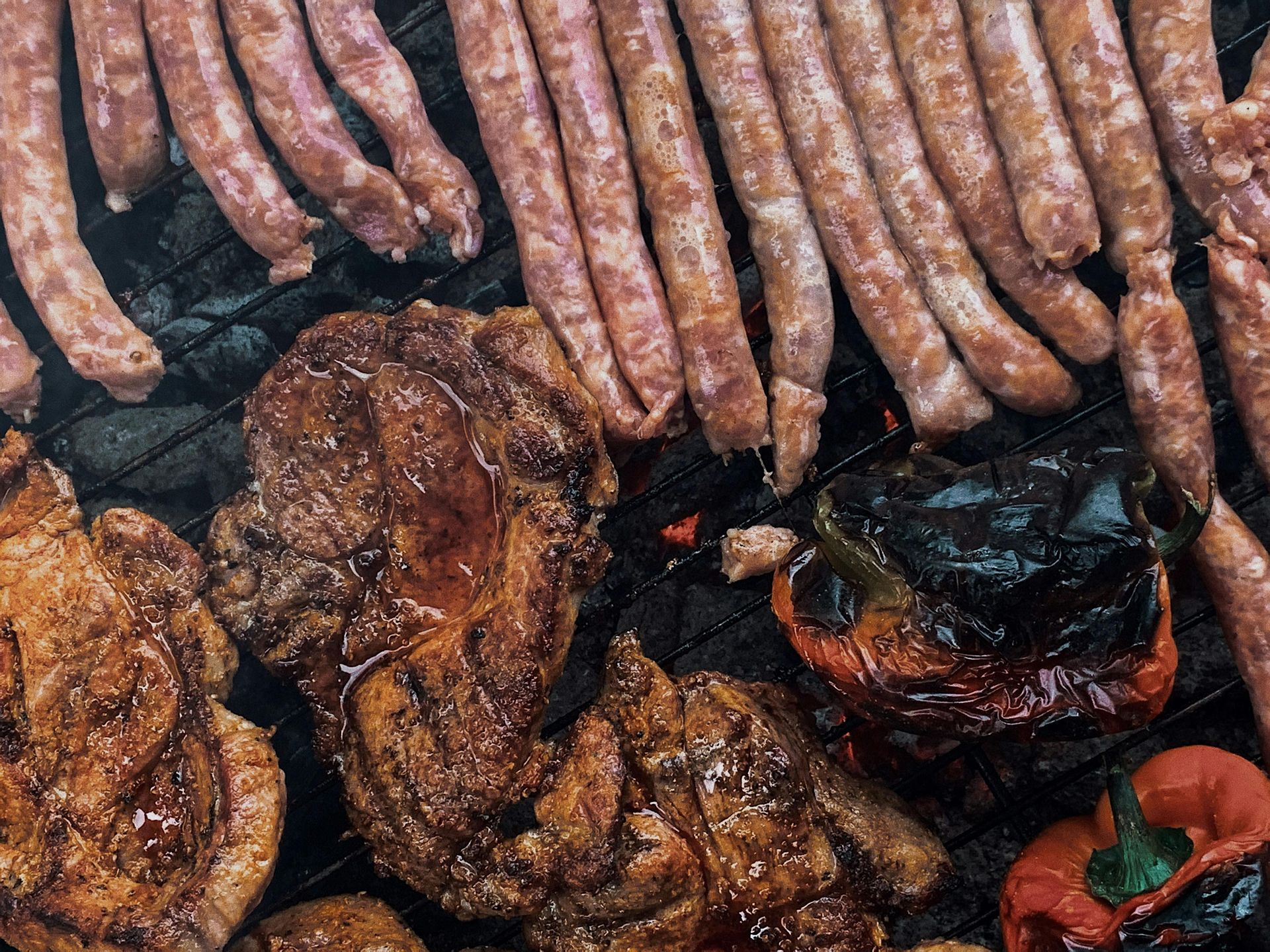 Grilled sausages and meat with a charred red pepper on a grill.