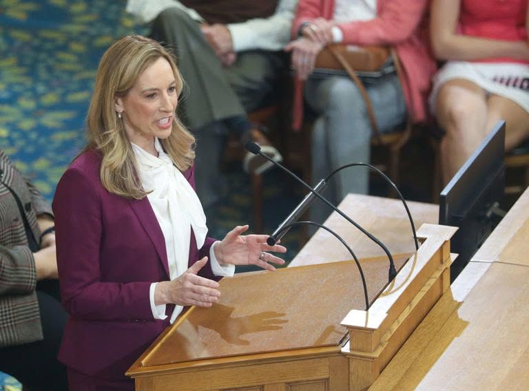 Woman in a burgundy blazer speaking at a podium in a hearing room, with microphones and a monitor nearby