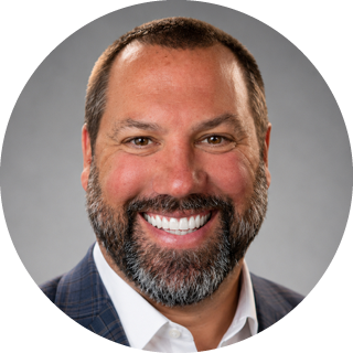 Smiling bearded man in a dark suit jacket and white shirt against a gray background
