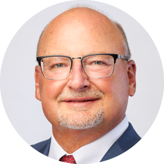 Headshot of a smiling man in glasses, wearing a blue suit and red tie, against a light gray background