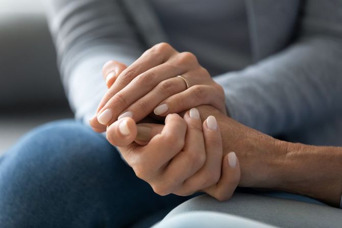 Two people gently holding hands on a couch, with one hand resting supportively over the other