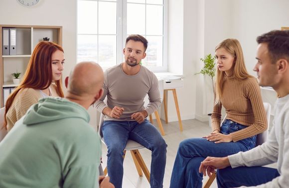 Five people in a seated group discussion in a bright living room, one person speaking to the others.