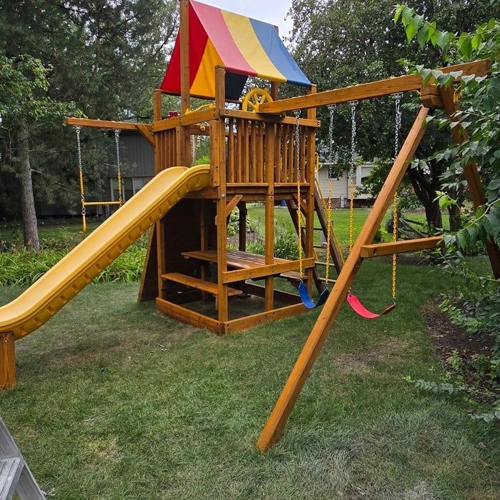 Wooden playground with slide, swings, and colorful roof, set in a grassy yard.