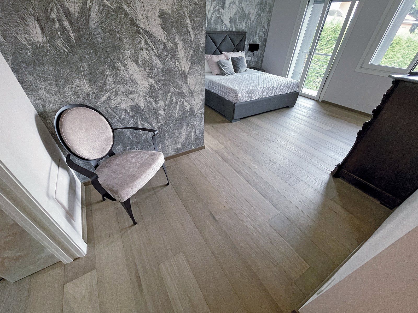 Camera da letto con parete grigia, sedia, letto e pavimento in legno. Luce intensa dalla finestra.
