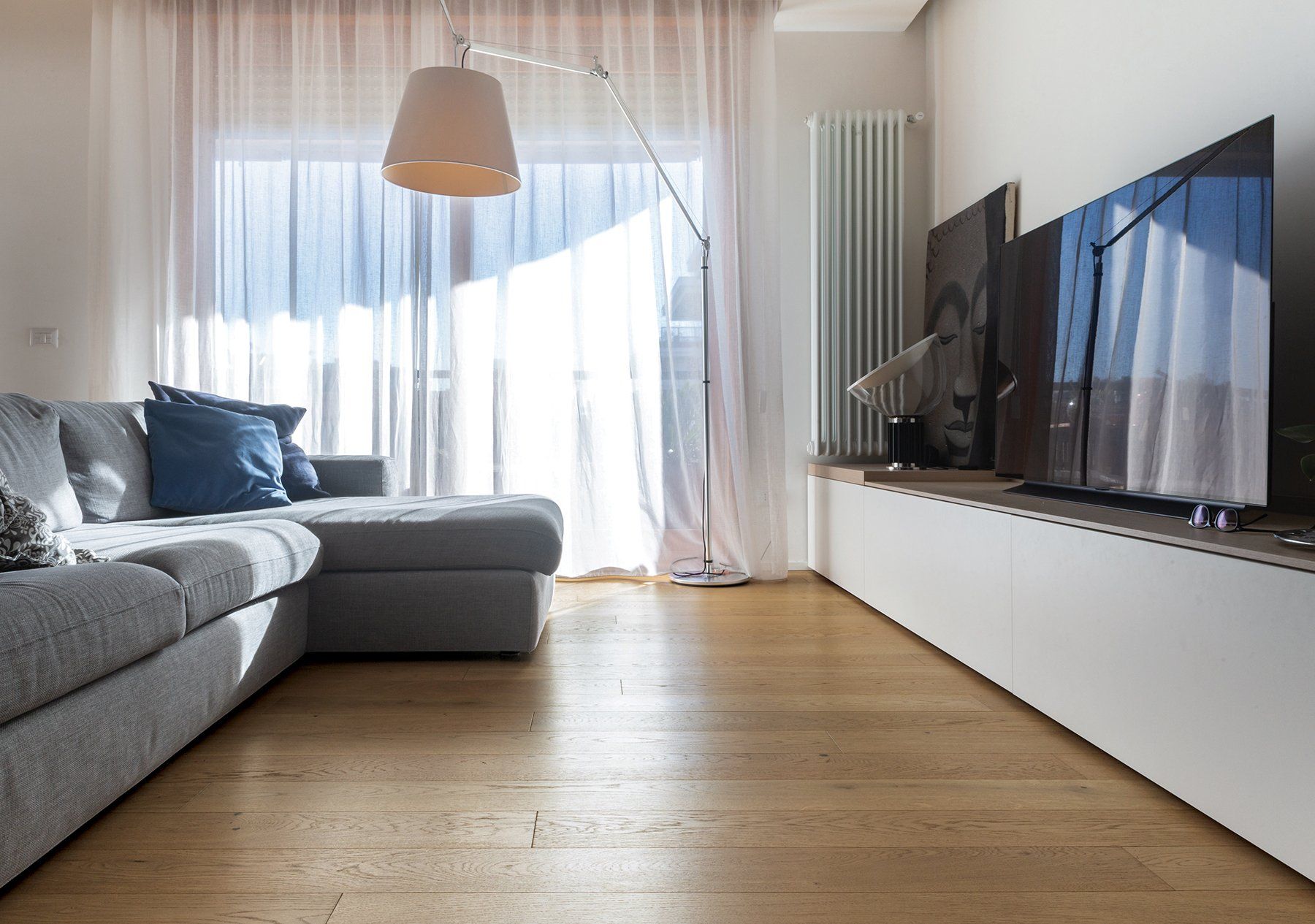 Soggiorno con divano grigio, grande TV, lampada ad arco e tende bianche trasparenti.