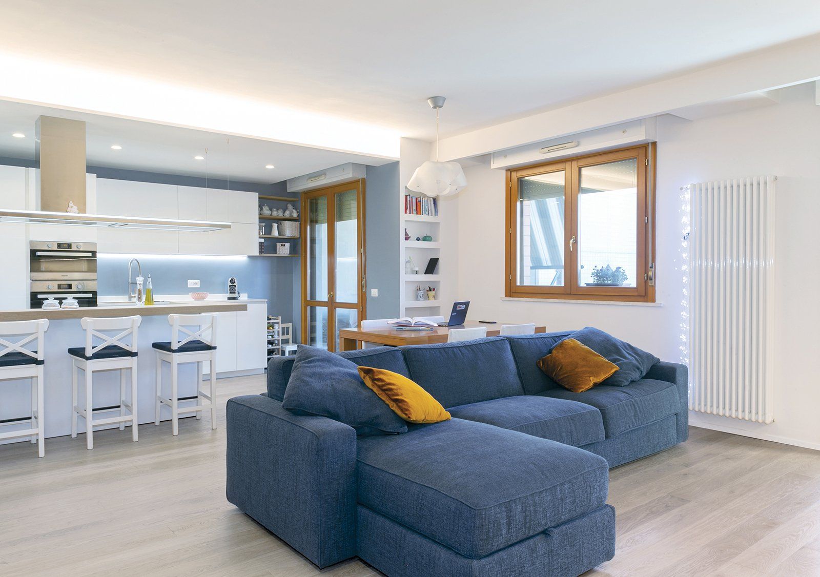 Soggiorno con divano componibile blu, cucina bianca e finestra in legno.